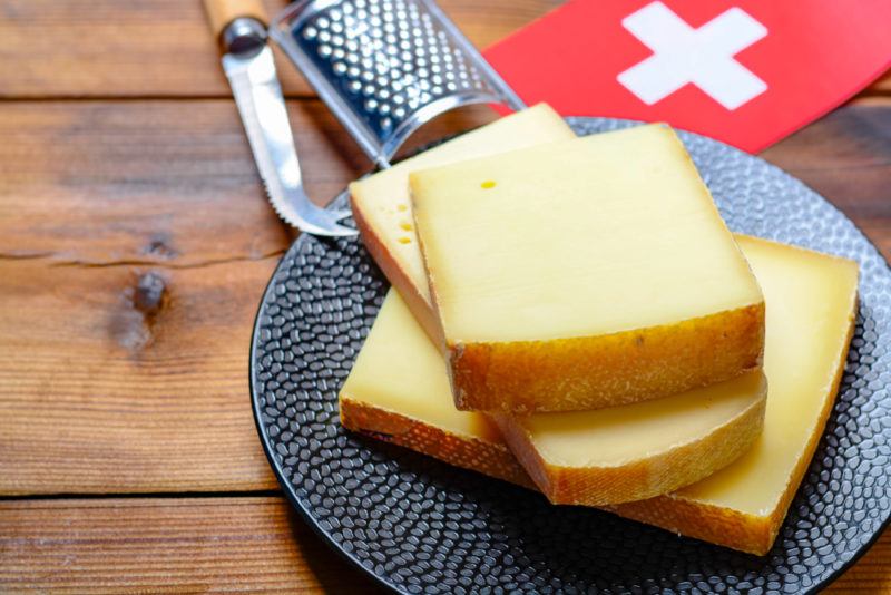 A gray plate or butting board, with harvarti cheese pieces. There is a small grater, a cheese knife and a Swiss flag in the background