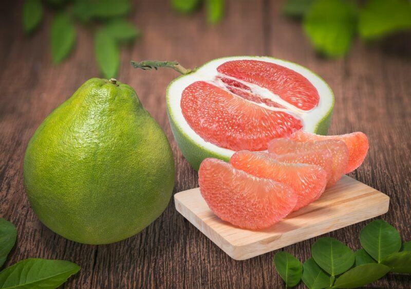 A wooden board with a single green pomelo, next to half a pomelo and some pomelo wedges