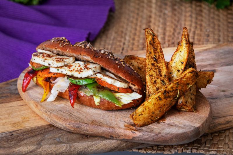 A wooden board on a wooden table, with a purple cloth in the background. On the board is a halloumi sandwich filled with grilled veggies, next to some potato wedges