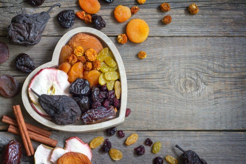 A wooden table with a white heart shaped bowl containing dried fruit and other pieces of dried fruit laid out across the table