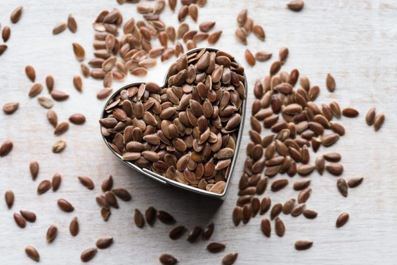 A heart shaped container of flaxseeds with more flaxeeds scattered on a table