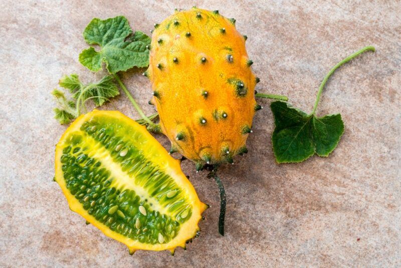 A whole horned melon, complete with spikes, next to half a horned melon, where the green interior pulp is visible