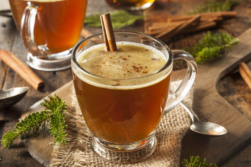 A glass mug of buttered rum next to Christmas trea leaves, with another mug in the background