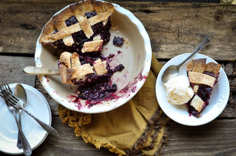 A deep dish of huckleberry pie with most pieces being gone, next to a small piece of the pie on a plate