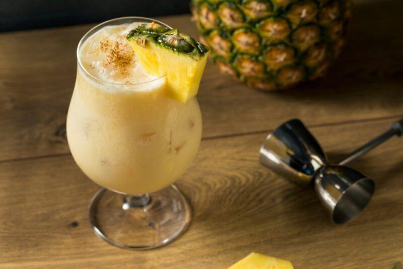 A wooden table with a painkiller cocktail in a hurricane glass, next to a cocktail jigger and a pineapple