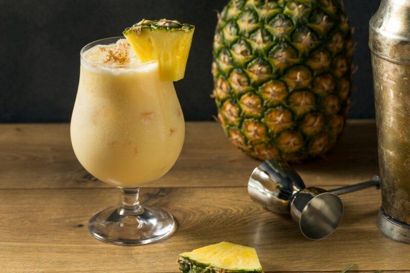 A hurricane glass containing a painkiller cocktail with a wedge of pineapple and a whole pineapple, next to a cocktail jigger