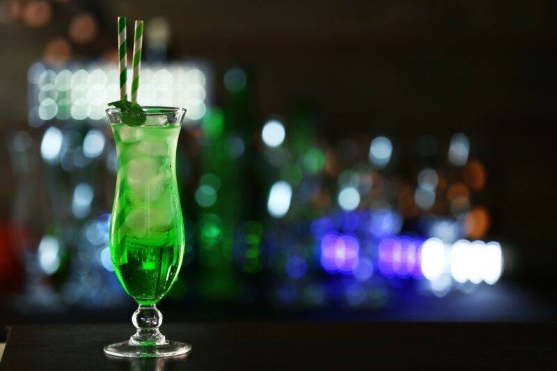 A tall hurricane glass containing a snake in the grass cocktail, in front of many out of focus bottles