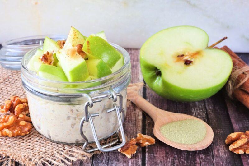 A small jar with overnight oats topped with apples, with half an apple and walnuts on the table