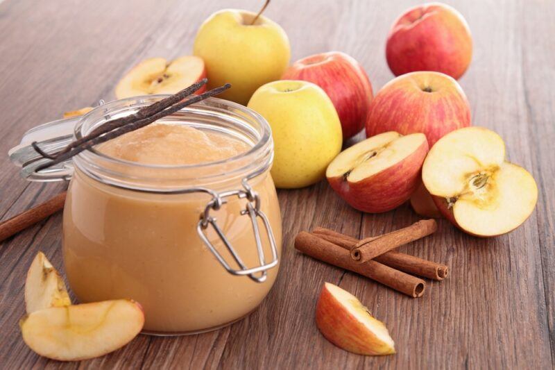 A jar of applesauce on a bench with cinnamon and many apples