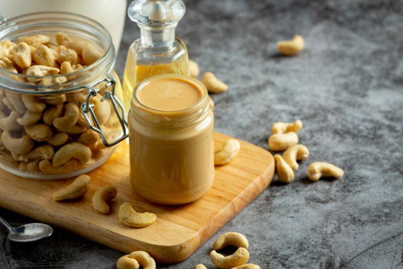 A wooden board with a container of cashew butter, some cashews, and some oil in the background