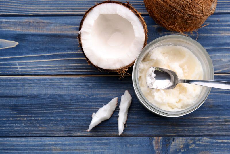 A top down image showing a jar of coconut oil with a spoon, next to a half coconut, a whole coconut, and some slivers on a blue table