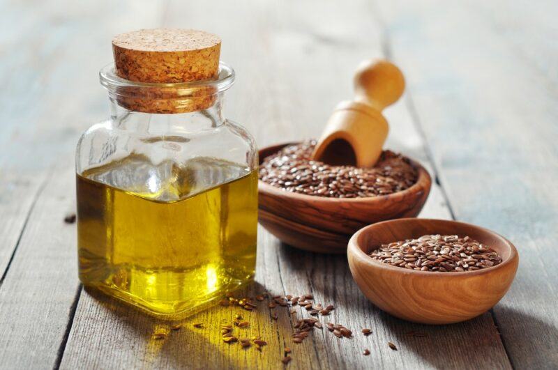 Two brown bowls with flax seeds, one that contains a scoop, next to a jar of flaxseed oil