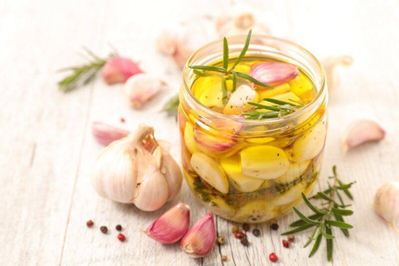 A light wooden table with a jar of garlic cloves, rosemary and olive oil, next to more garlic cloves on the table