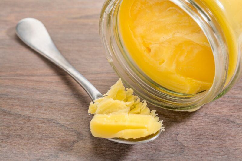 A jar of ghee on its side, next to a spoon filled with ghee on a wooden table