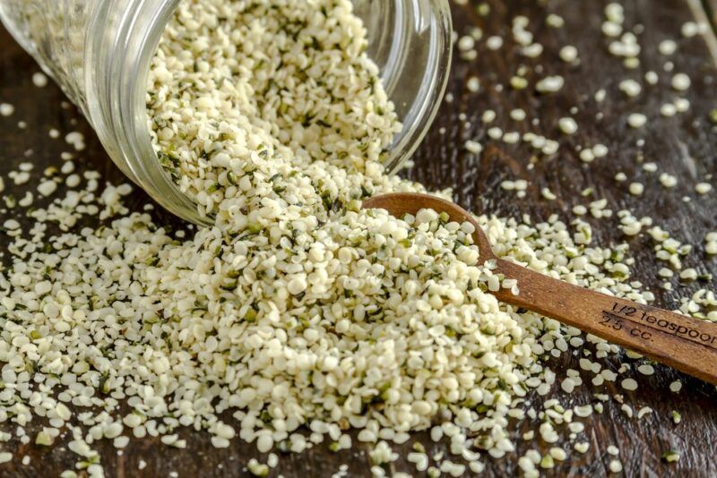 A jar of hemp hearts that is being tipped out onto a wooden table, with a small spoon in the midst of it