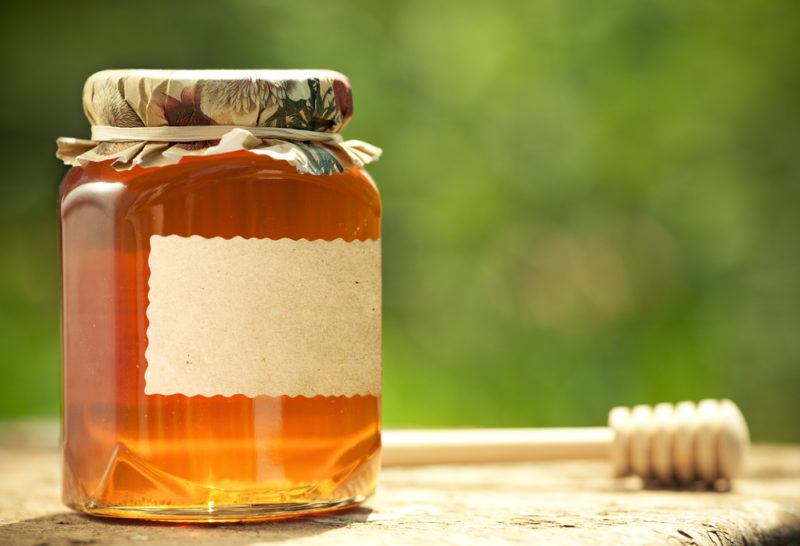 A jar of honey next to a honey stirrer against a green background