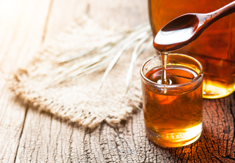 A jar of maple syrup and a spoon, in front of a bottle of maple syrup and next to a cloth