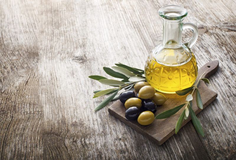 A jar of olive oil, with olives on a plate on a wooden table