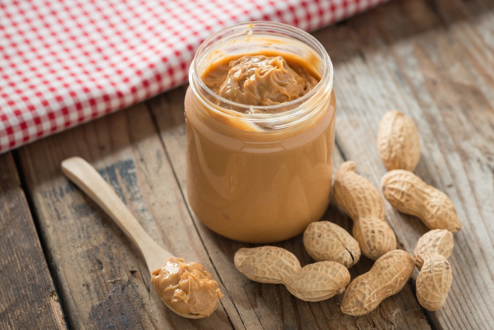 A wooden table with a jar of peanut butter, peanuts in their shells, and peanut butter on a spoon