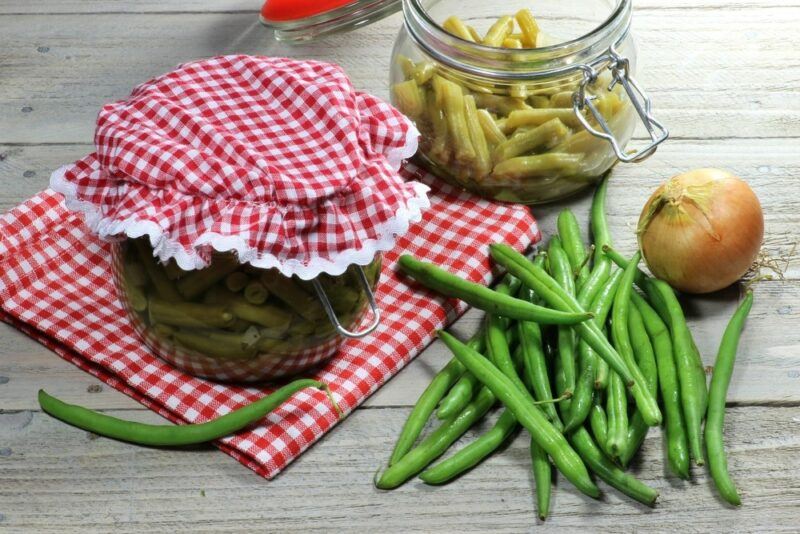 A red and white checkered cloth, with a jar of pickled beans, next to plenty of fresh beans and a jar that's being prepared in the background