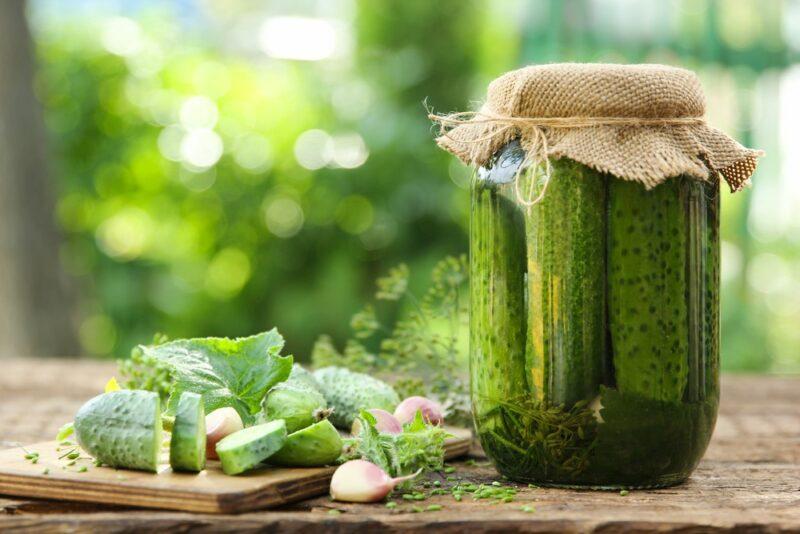 A glass jar of pickles with brown cloth on top, next to some sliced pickles