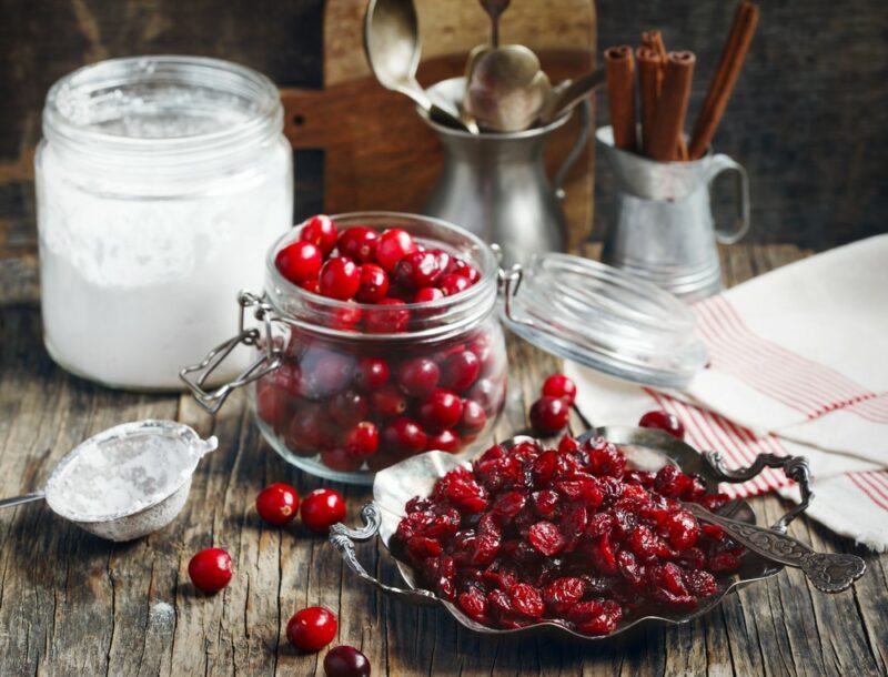 A jar of powdered sugar on a table, with a jar of fresh cranberries in front of it and a jar of dried cranberries in front of that