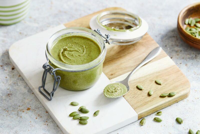 A wooden board with pumpkin seed butter, a spoon, and some pumpkin seeds scattered around