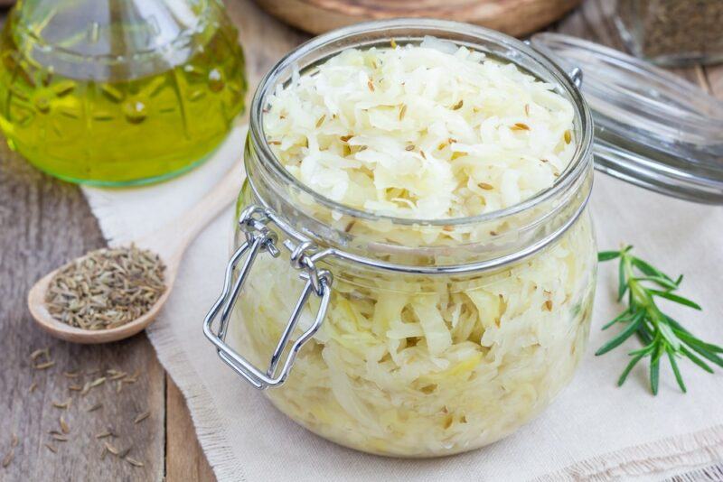 A large glass jar of sauerkraut on a table. There is a wooden spoon of caraway seeds next to it, plus a glass container of oil further in the background.