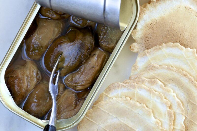 A can of smoked oysters opened on a table with a fork, next to some plain crackers