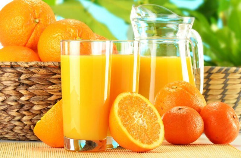 Two large glasses of orange juice and a jug of the juice, next to a basket of oranges and some oranges on the table