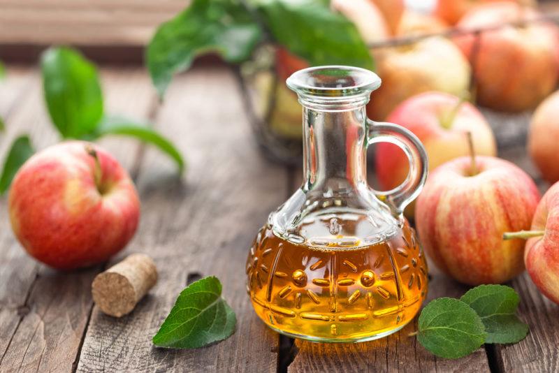 A small glass jug of apple cider vinegar next to a selection of red apples on a table