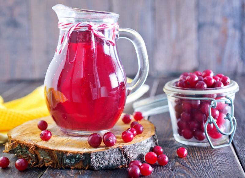 A jug of cranberry juice on a wooden table, next to a glass jar of cranberries with more cranberries scattered on the table