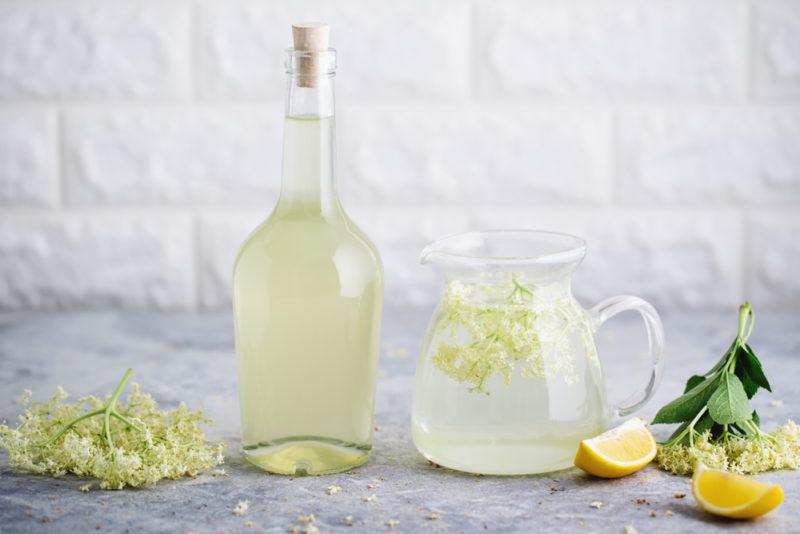 A bottle of elderflower cordial and a jug of the cordial, with elderflowers and lemons on the table