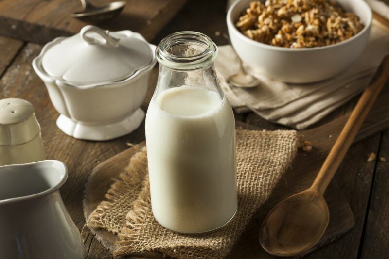 A glass bottle of milk on a table with other items for breakfast, including a bowl of cereal, sugar, and a spoon