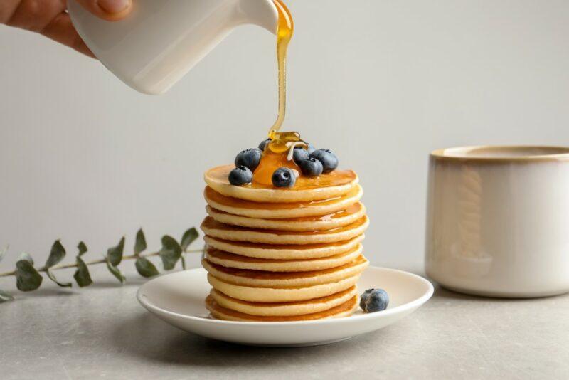 A stack of pancakes on a white table with berries on top, where someone is pouring honey onto the pancakes