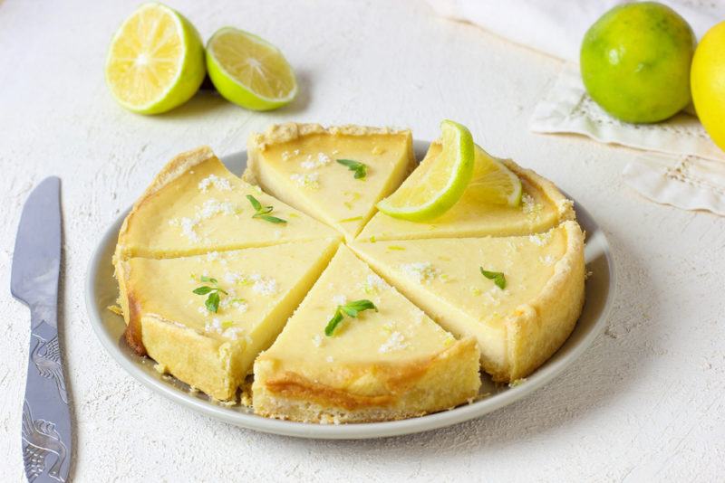 A white plate containing a key lime pie that has been cut into segments, next to a knife and some limes