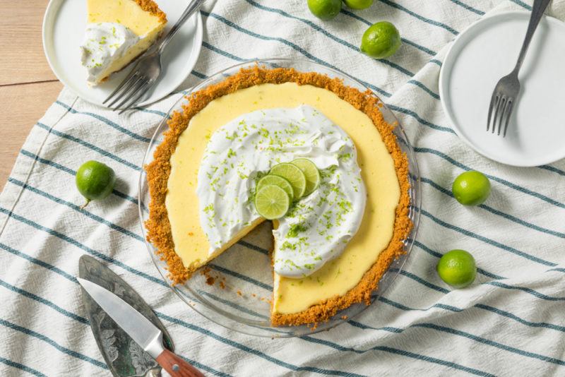 A top-down view of a key lime pie with a small piece cut out of it and on a separate plate. The pie is on a tablecloth, with limes and cutlery scattered around