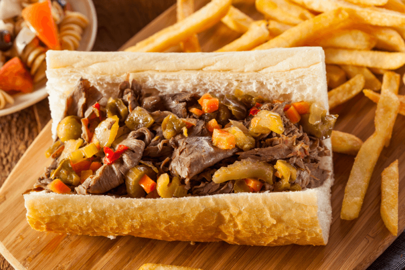 A large roll containing an Italian beef sandwich on a wooden board with chips in the background.