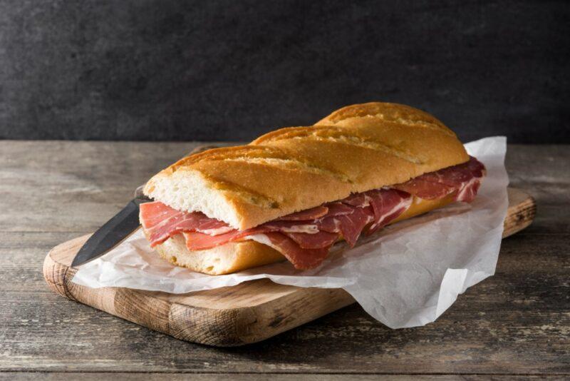 A large Spanish sandwich filled with meat on a piece of white cloth or paper. The sandwich is known as a bocadillo