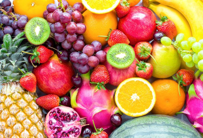 A flat lay of many different types of fresh fruit, including grapes, kiwis, pineapples, pomegranates, and oranges