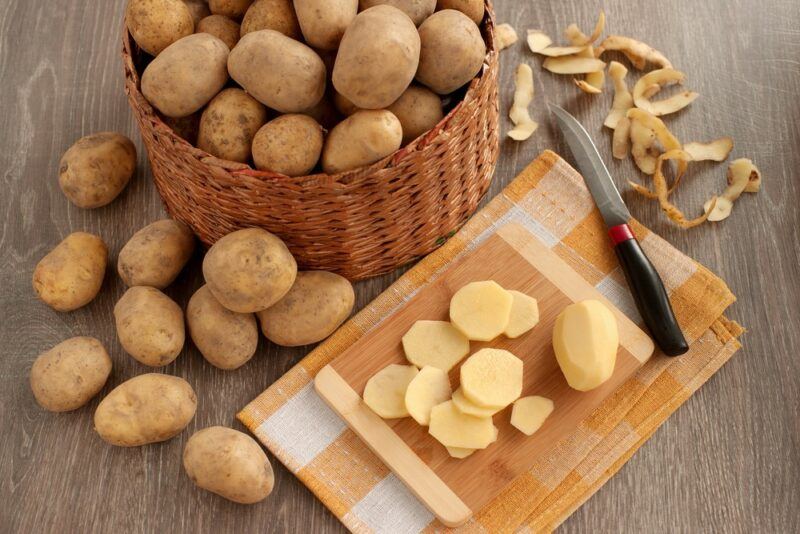 A large basket of potatoes on a table, with some more scattered around and sliced potatoes on a board