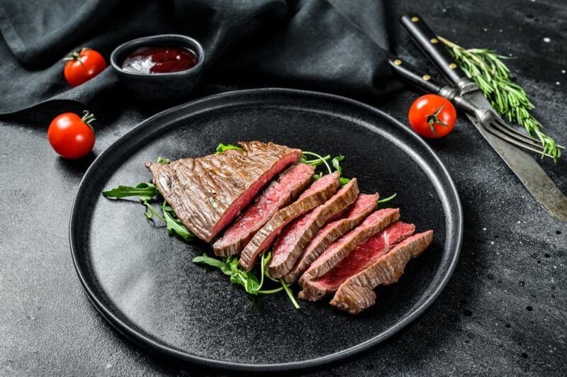 A large black plate with a flat iron steak that has been sliced into thin pieces, surrounded by rosemary. There is more rosemary, some sauce, and some cherry tomatoes on the table