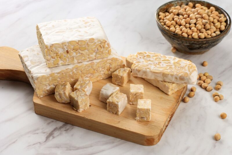 A wooden tray with a tempeh cake that's been cut into smaller pieces, next to a bowl of soybeans