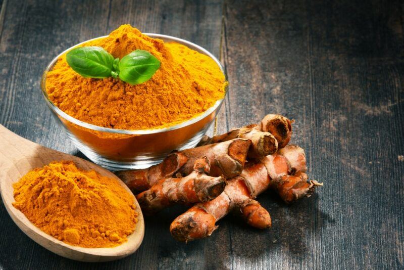 A dark table with a bowl of turmeric powder, a large spoon of the powder, and some turmeric roots