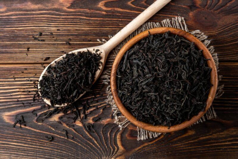 A large wooden bowl of black tea leaves, with a spoon filled with more tea leaves