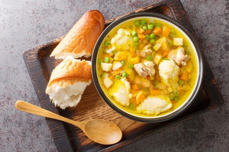 A wooden tray with a large bowl of chicken dumpling soup, a spoon, plus some pieces of crusty bread