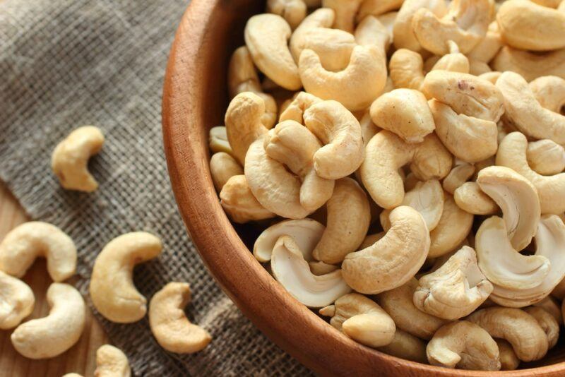 A large brown bowl with raw cashews next to some cashews on the table