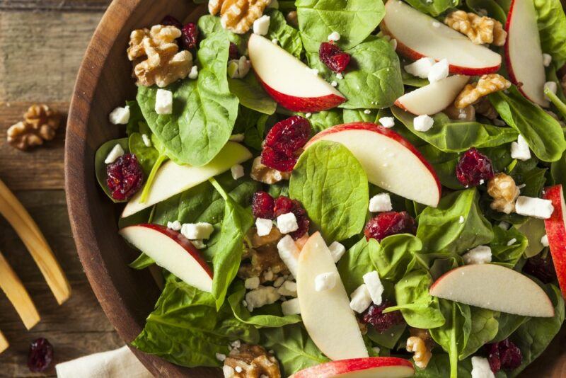 A salad in a large brown bowl, which include spinach, feta, walnuts, apples, and dried cranberries