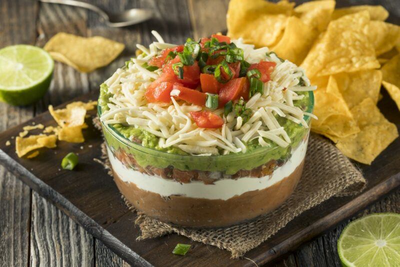 A large glass dish with a seven layer dip that's been topped with cheese. There are tortilla chips all around it