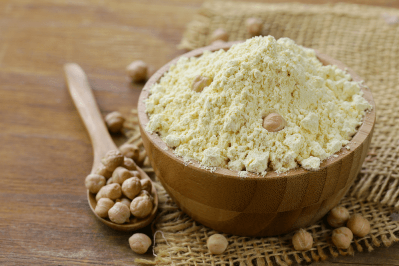 A wooden table with a wooden spoon containing chickpeas, plus a large wooden bowl of chickpea flour